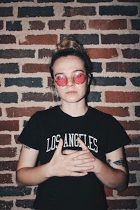 a woman wearing sunglasses and a black t - shirt in front of a brick wall